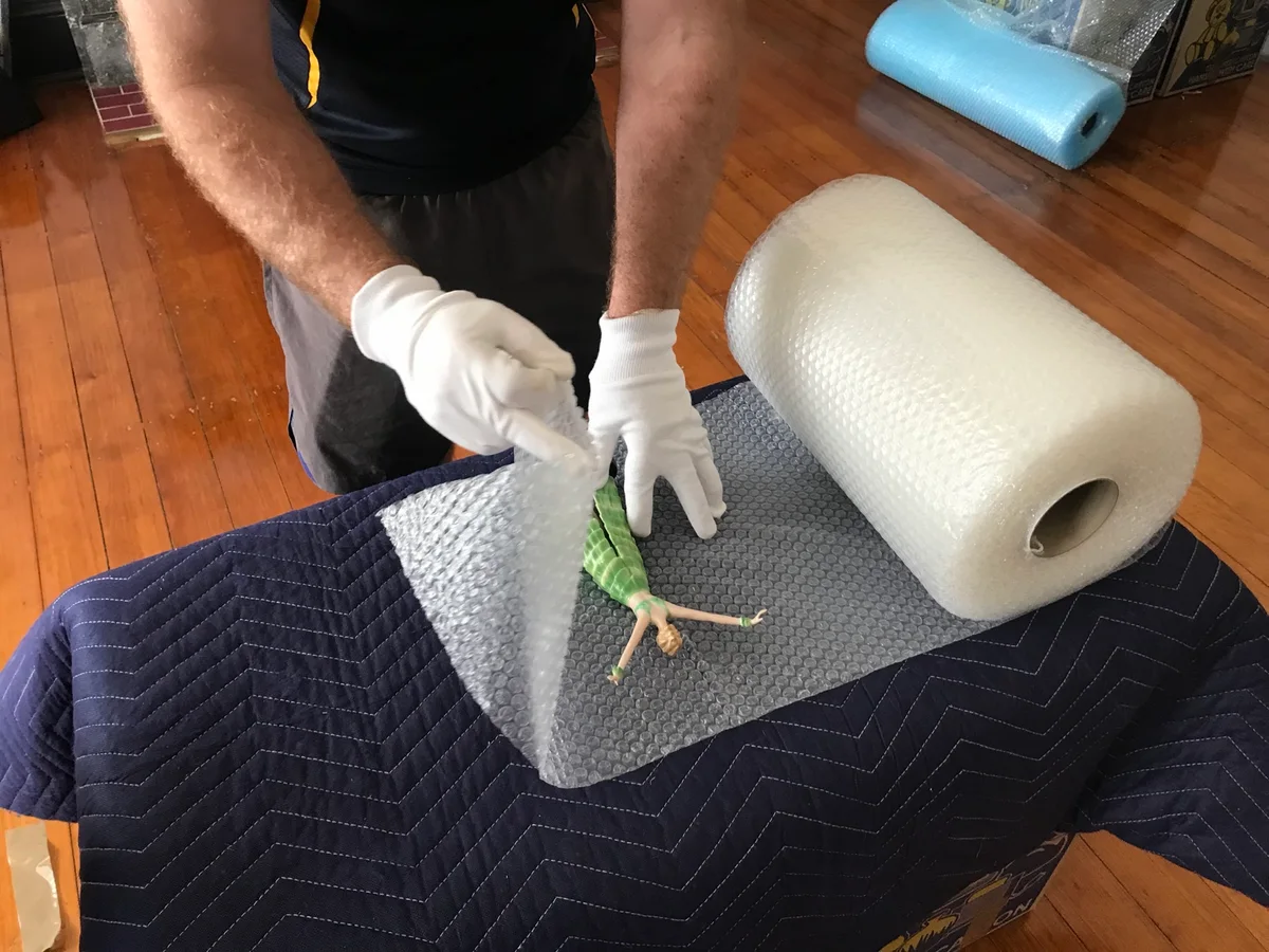Gloved hands wrapping a delicate figurine in bubble wrap on a protective moving blanket