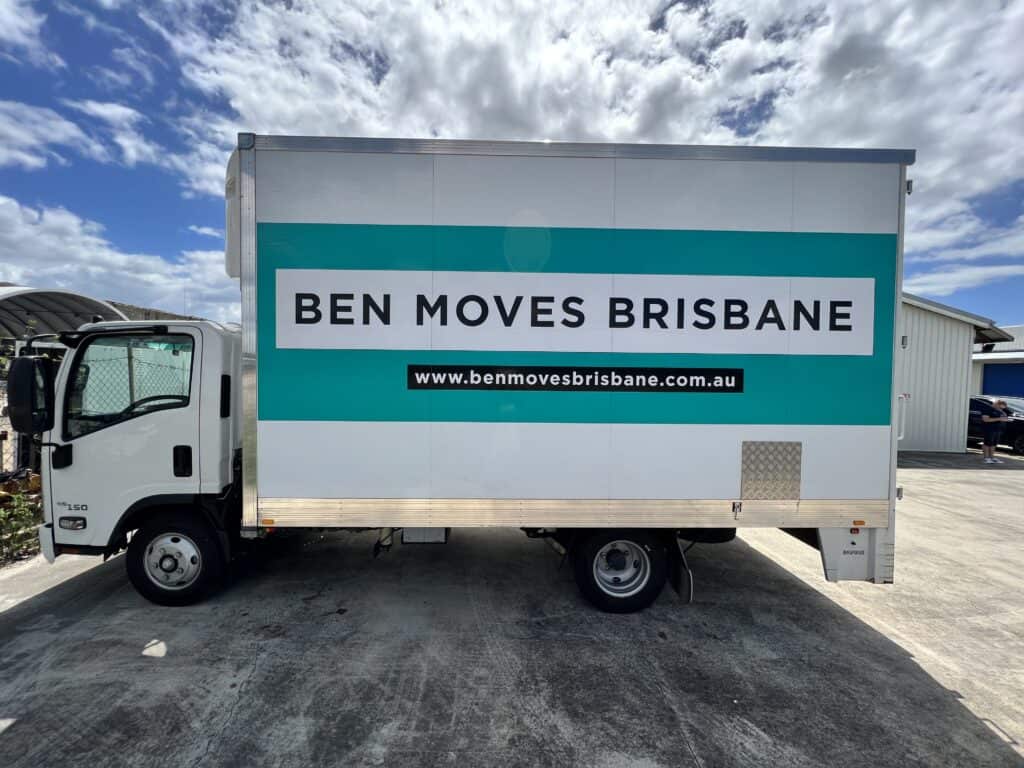 A small moving truck parked on concrete in front of sheds and blue skies with clouds in the background